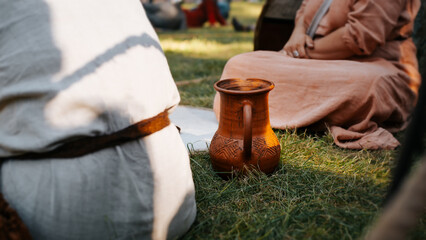 Rustic ceramic antique jug standing near people in ancient clothes on green grass outdoors. Medieval concept. Close-up, low angle view