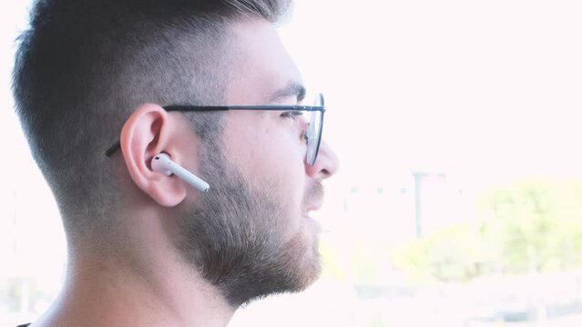Using ear buds, young brown haired teen man using ear buds. Talking to phone by wearing wireless ear buds. Speaking and listening. Over shoulder view footage of young male using in-ear headphones.