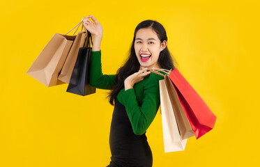 Young asian in green long-sleeved shirt and black dress carrying paper shopping bags posing smiling and happy on the yellow screen background. Shopping woman lifestyle.