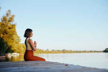 Yogi woman practices breathing exercises on the dock while holding hands in a namaste pose.