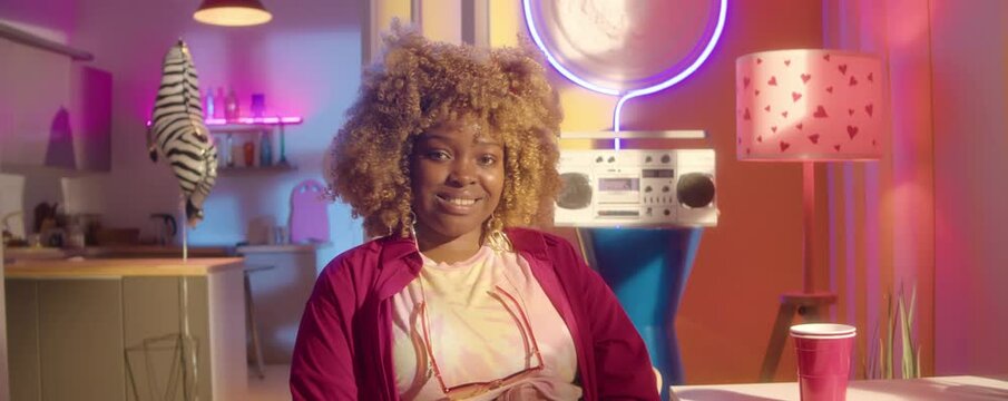 Zoom in portrait shot of young African American girl in bright outfit sitting in party room with neon decorations and smiling on camera
