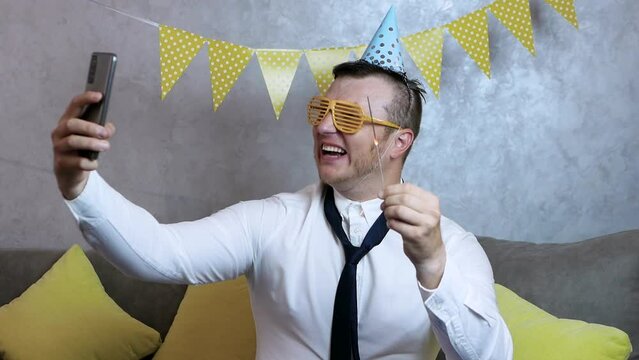 Loud smiling man happy birthday. Very happy young man celebrates his birthday, a man with begal lights in his hands, holiday garlands. Handsome young man in a festive hat on his head takes a selfie.