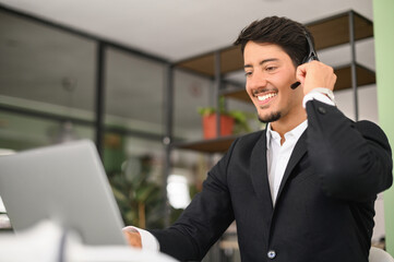 Portrait of confident latin man in wireless headset using laptop computer sitting in contemporary office space, male employee is making video call, talking online on distance