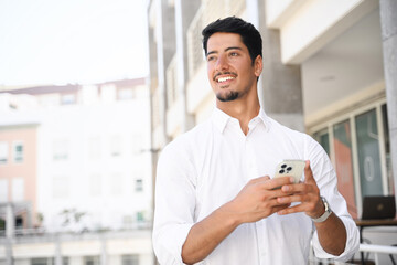 Happy and smiling latin dark-haired man in white shirt using smartphone outdoors, texting, chatting...