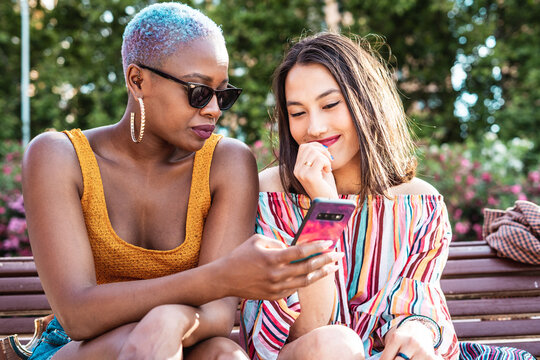 Diverse girlfriends using smartphone on bench