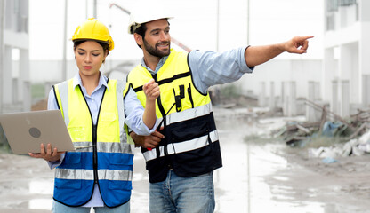 Engineer man and female architect wear safety helmets discuss housing development project at construction site using laptop computer. Contractor manager examining building estate infrastructure.