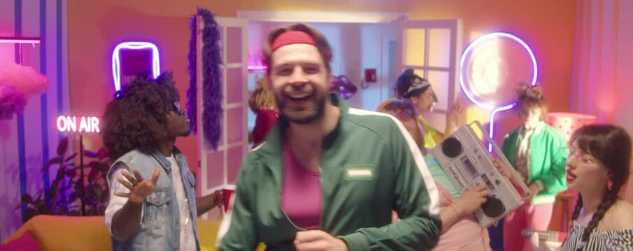 Zoom in shot of happy bearded man wearing green green tracksuit and fitness headband looking and camera and dancing energetically at 90s disco party with friends in room with neon light