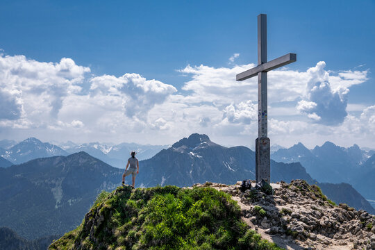 Woman standing on peak next to cross looking at mountain, Tegelberg, Germany