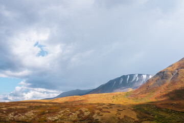 Motley autumn landscape with sunlit hills and mountain range silhouette under dramatic cloudy sky. Vivid autumn colors in mountains. Sunlight on multicolor hills and rainy clouds in changeable weather