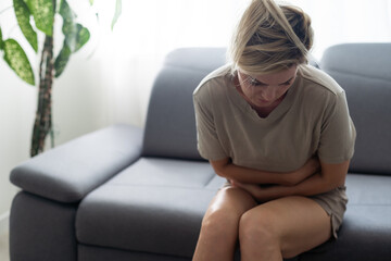Sad, upset, unhappy woman holding hands on stomach suffering from abdominal pain with close eyes, having menstrual period, food poisoning, gastritis, diarrhea, feeling unwell sitting in livingroom.