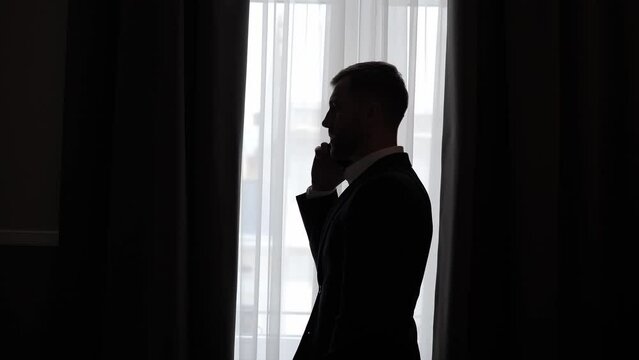 Silhouette of a business man standing near window talking on a mobile phone having phone conversation. Successful confident businessman making smartphone call. Private safety communication indoor.