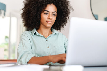 Concentrated young businesswoman in casual wear is looking at the laptop screen, solving tasks, freelance woman working remotely from home office