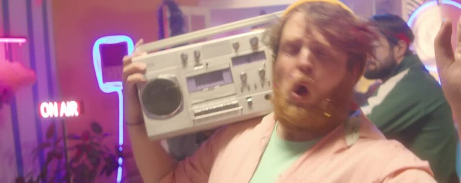 Young man with painted nails and confetti on beard dancing with ghetto blaster on shoulder at 90s party with friends in room colorful disco lights