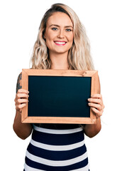 Young blonde woman holding blackboard smiling with a happy and cool smile on face. showing teeth.