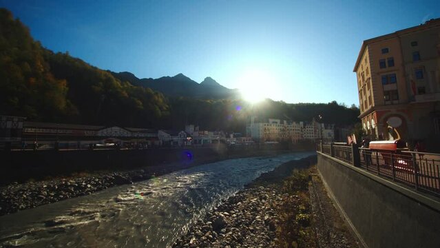 Embankment with hotels and restaurants of olympic village rosa khutor with flowing water stream. tourists are walking along the river. sun is shining bright.