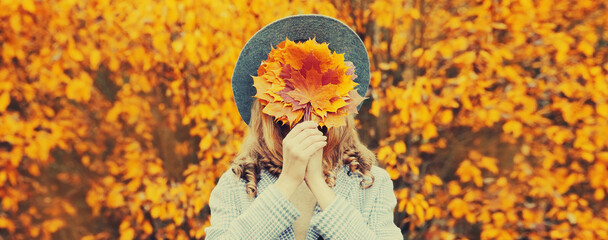 Autumn portrait of stylish woman covering her face with yellow maple leaves wearing round hat in the park