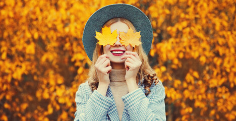 Autumn portrait of beautiful happy smiling woman with yellow maple leaves wearing round hat in the...