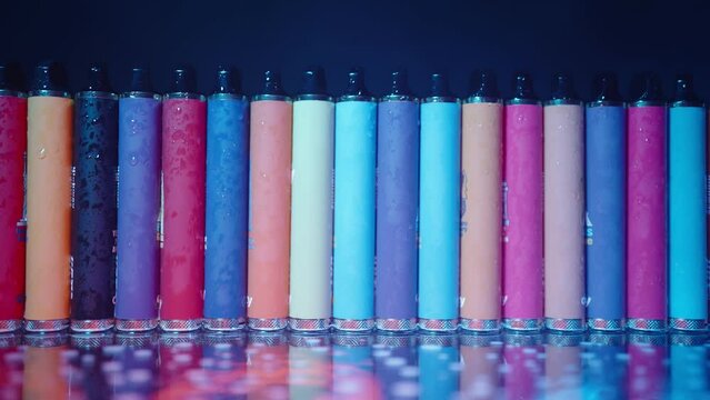 Lots of E-Cigarettes and Vapes with Smoke in Neon Lighting and Light from an Ambulance Flasher. Concept of Bad Habits. Modern Smoking Electronic Cigarettes. Slow Motion
