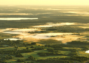 Fototapeta premium Foggy sunrise. Nature at dawn in fog, aerial view. Morning mist haze in agricultural field. Sunny morning scene in misty in rural. Misty landscape. Sunny foggy hills on sundown. Dawn in hillside.