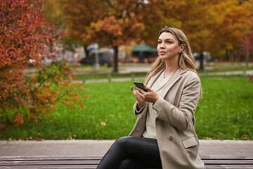 a beautiful woman against the background of a red autumn bush in a light coat sits on a bench and holds a smartphone in her hands. yellow trees and fallen leaves.