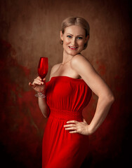 A woman in a red dress stands on a brown background and holds a wine glass in her hand. Middle aged blonde. The woman has a short haircut.