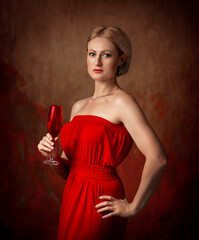 A woman in a red dress stands on a brown background and holds a wine glass in her hand. Middle aged blonde. The woman has a short haircut.