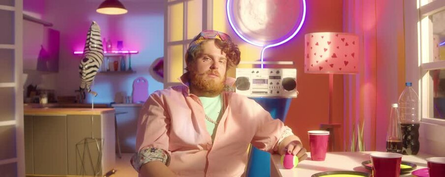Zoom in portrait shot of bearded man with painted nails in party outfit sitting in brightly colored room with neon decorations and posing for camera with confidence