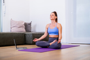 Fototapeta na wymiar Calm woman dressed in sportswear, meditates on the floor in her living room, listening to spiritual practices classes on laptop, poses in lotus pose, tries to relax. Yoga concept