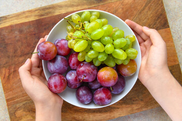 women's hands with a plate full of green grapes and plums on a wooden board, the concept of fresh fruit and healthy food