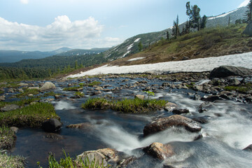 Mountain stream during spring flood, water breaks against rocks. Rivulet in mountainous area, rock ridge on horizon. Glaciers and snow on hillsides. Clouds over riverbed. - 525601019
