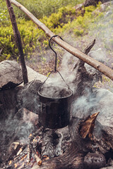 Tourist pot hangs over campfire. Survival in wild for cooking. Organization of hearth of stones for heating. - 525601024