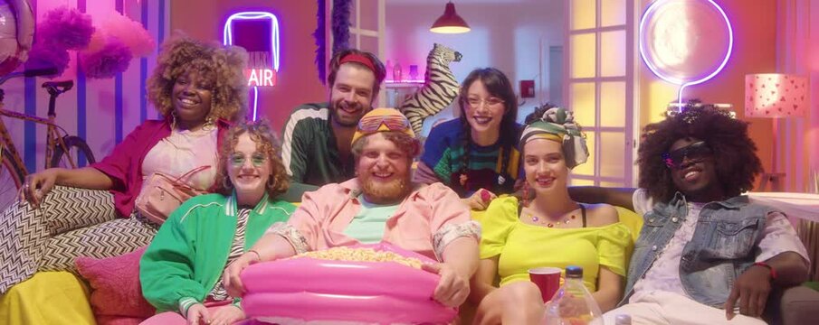 Zoom in shot of joyous diverse friends in trendy bright outfits sitting on sofa with popcorn and drinks, looking at camera and laughing while watching comedy movie in room with neon light