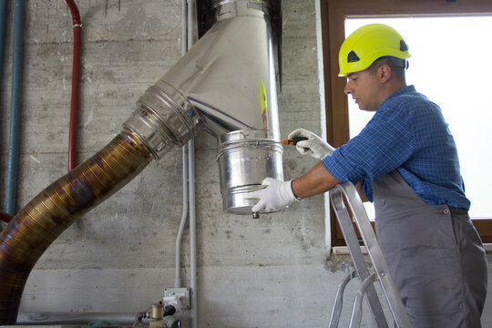 Image of a hard-hatted handyman plumber on a ladder repairing and assembling a stainless steel pipe of a boiler flue. Do it yourself work. 