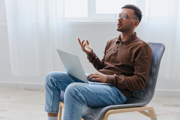 Thoughtful pensive serious young man in eyewear raise hand up thinking about best solution for project sitting in chair at home. Remote Job Distant Work Communication Modern Profession concept