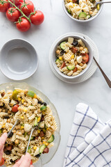 Hands holding serving utensils to serve up bowls of fresh homemade pasta salad.