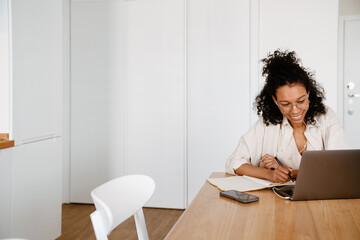Black woman writing down notes while working with laptop at home
