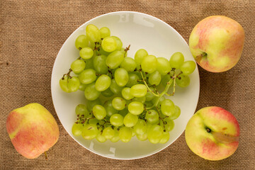 One bunch of white grapes and three apples with a ceramic plate on burlap, close-up, top view.