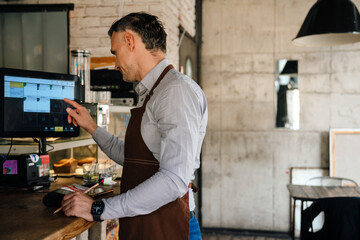Adult white waiter wearing apron using computer while working in cafe