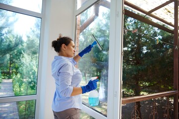 Multi-ethnic maid doing household chores wiping windows, spraying detergent and using scraper...