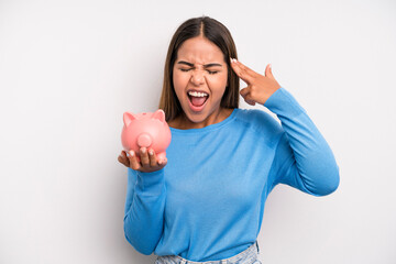 hispanic pretty woman looking unhappy and stressed, suicide gesture making gun sign. piggy bank...