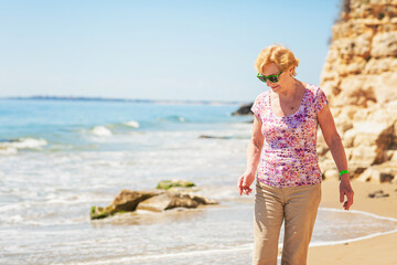 Senior woman in sunglasses stands on the beach and looks at the sea. Old woman enjoying life near the sea