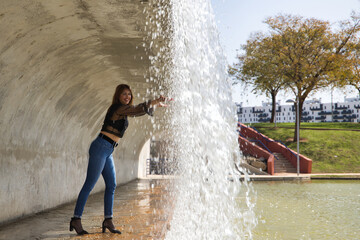 Attractive mature woman in transparent black shirt, jeans and heels, touching water falling from a waterfall. Concept maturity, beauty, fashion, water, happiness.