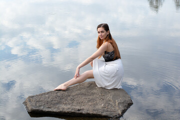 a girl in a dress on a stone in the river with a reflection of the sky with clouds in the water