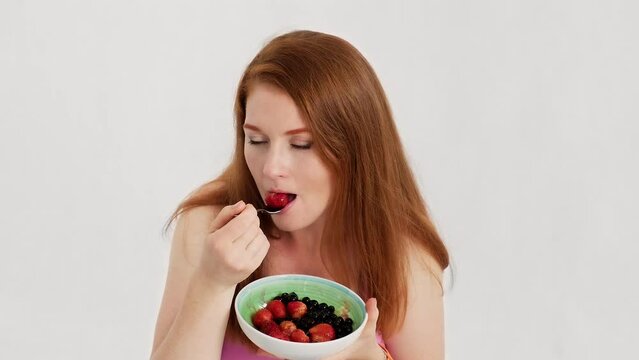 Fit woman eat strawberry fruit in studio at white background. Redhead woman eating strawberries with pleasure. Healthy nutrition. Organic dessert. 4K, UHD