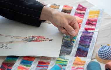 Selective focus on hands of fashion designer comparing or choosing color of threads with color of fashion clothes on sketch paper, with fabric and threads color chart and tailor tools on working desk