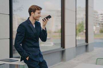 Confident businessman with smartphone using voice assistant app while leaning on cafe table outdoors
