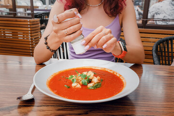 A woman adds salt from a saltcellar to soup in a restaurant. An overabundance of consumption of this seasoning can lead to cardiovascular diseases and gout - obrazy, fototapety, plakaty