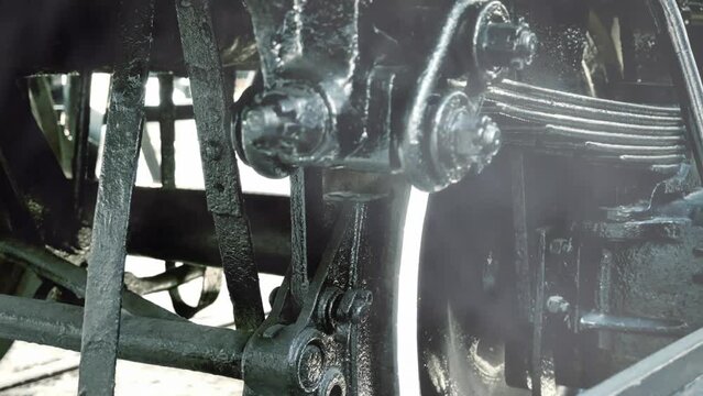 Closeup detail shot of the wheel of an old train, showing the mechanisms of a black iron giant ready to move, with white steam rising from below.

