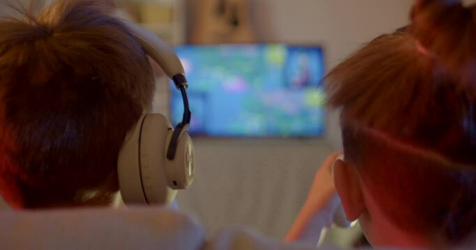 Backs heads of teenagers play the game. Against background of the monitor in bokeh. Controllers in hand. Leisure activities in the evening at home without parents. Headphones on the head.