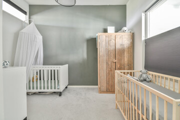 Interior of white room with baby crib and armchair near window with blue chest of drawers in...